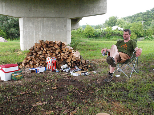 Front view of a person sitting in a camp chair, in a field under a cement bridge, next to boxes of beer and a log pile